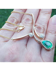 Hand holding a yellow gold necklace with a natural emerald gemstone pendant against a blurred green background