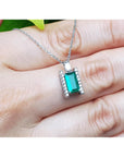 Baguette cut emerald gemstone pendant on a white gold chain held between fingers.