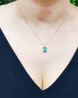 A woman wearing a necklace with a natural baguette cut emerald on a plain natural background.