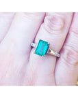 18K gold ring with a deep bluish-green emerald and tapered baguette side diamonds on a hand.