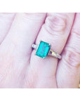 18K gold ring with a deep bluish-green emerald and tapered baguette side diamonds on a hand.