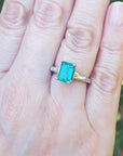 Ring with an emerald and baguette side diamond ring on a person's finger against a blurred green leafy background.
