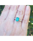Ring with an emerald and baguette side diamond ring on a person's finger against a blurred green leafy background.