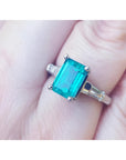 A three stone genuine emerald and baguette side diamond ring in white gold on a finger.