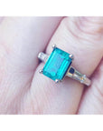 A three stone genuine emerald and baguette side diamond ring in white gold on a finger.