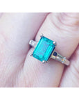 A three stone genuine emerald and baguette side diamond ring in white gold on a finger.