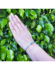 Colombian emerald and diamond bangle bracelet in 14K white gold on a wrist against green leaves.