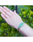 Hand wearing a natural emeralds and diamonds bangle bracelet with a green leafy background.