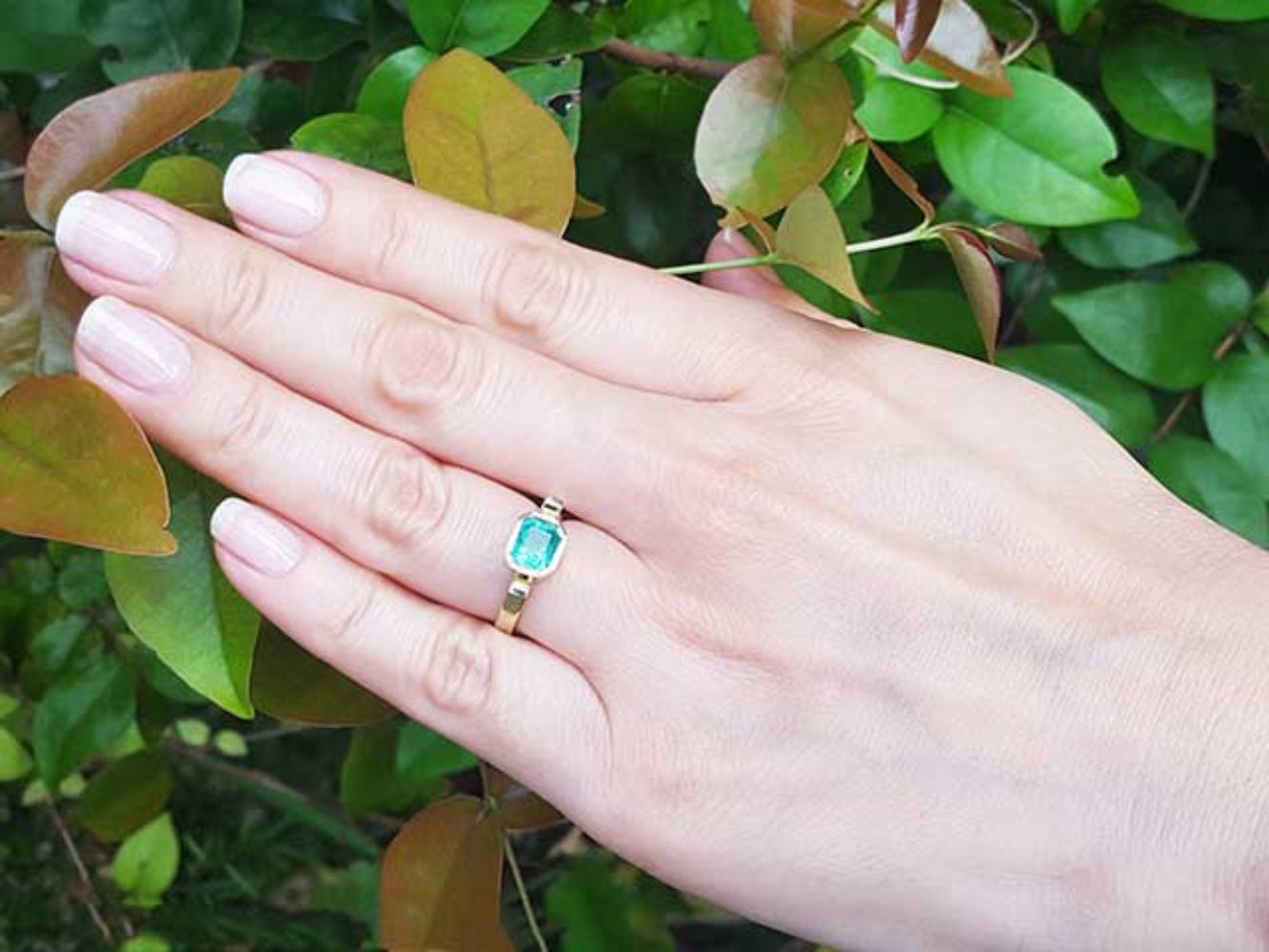Hand wearing a colombian emerald solitaire ring bezel set against a green leafy background