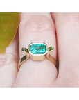 Bezel-set emerald cut Colombian emerald ring in 14K yellow gold on a finger.