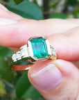 A natural Colombian emerald ring for women held between fingers with a blurred green leafy background.