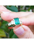 A natural Colombian emerald ring for women held between fingers with a blurred green leafy background.