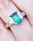 A bezel set emerald ring with accent princess cut diamonds on a finger.