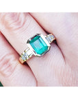 A bezel set emerald ring with accent princess cut diamonds on a finger.