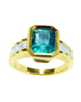 A women's bezel set emerald ring with accent princess cut diamonds on a top profile view position.
