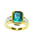 A women's bezel set emerald ring with accent princess cut diamonds on a top profile view position.