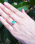 Hand wearing a bezel set emerald ring with accent princess cut diamonds against a leafy background.