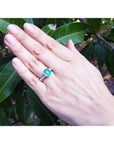 Hand wearing a bezel set emerald ring with accent princess cut diamonds against a leafy background.