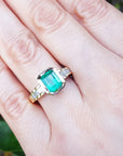 A May's birthstone brides' emerald engagement ring on a hand against a blurred natural background.