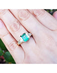 A May's birthstone brides' emerald engagement ring on a hand against a blurred natural background.