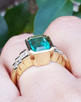A women's Colombian emerald ring bezel set on a finger against a blurred green leafy background.