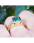 A women's Colombian emerald ring bezel set on a finger against a blurred green leafy background.