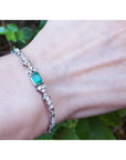 A white gold bracelet with a center natural emerald on a wrist against a natural background.