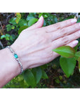 Hand wearing a bracelet with emerald and diamonds against a green leafy background.