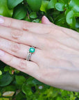 Hand wearing a ring with an emerald stone against a green leafy background