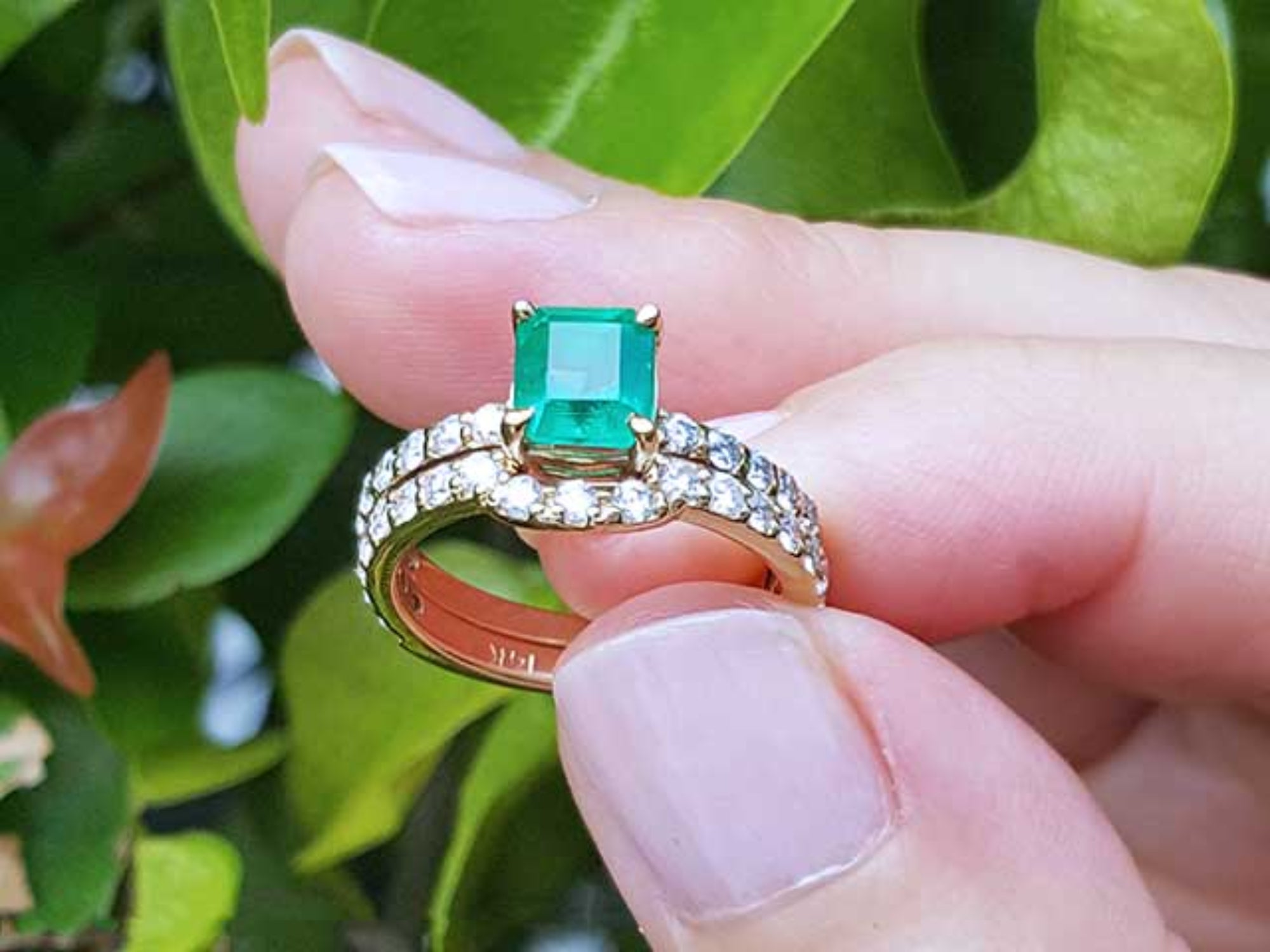 An emerald ring with matching band set held between fingers against a green leafy background.