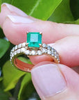 An emerald ring with matching band set held between fingers against a green leafy background.