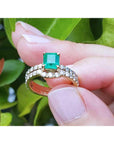 An emerald ring with matching band set held between fingers against a green leafy background.