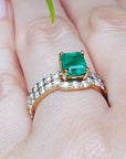 A yellow gold emerald bridal ring with the matching band set on a finger against a blurred natural background.