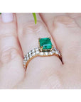 A yellow gold emerald bridal ring with the matching band set on a finger against a blurred natural background.