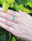 Hand displaying a circle of life pendant with an oval cut emerald against a green leafy background.