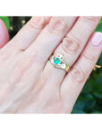 Hand wearing an emerald claddagh ring crafted in 14k yellow gold against a blurred green background.
