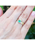 Hand wearing an emerald claddagh ring crafted in 14k yellow gold against a blurred green background.