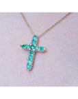 Cross-shaped necklace with natural emeralds on a light pink skin background.