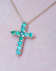 Cross-shaped necklace with natural emeralds on a light pink skin background.