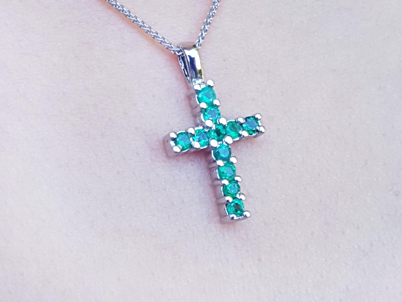 A white gold cross pendant with round cut natural emeralds on a light pink skin background.