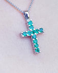 A white gold cross pendant with round cut natural emeralds on a light pink skin background.