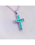 18K white gold cross pendant with round-cut emeralds on a delicate chain, worn on a person.
