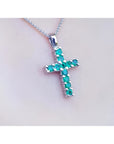 18K white gold cross pendant with round-cut emeralds on a delicate chain, worn on a person.