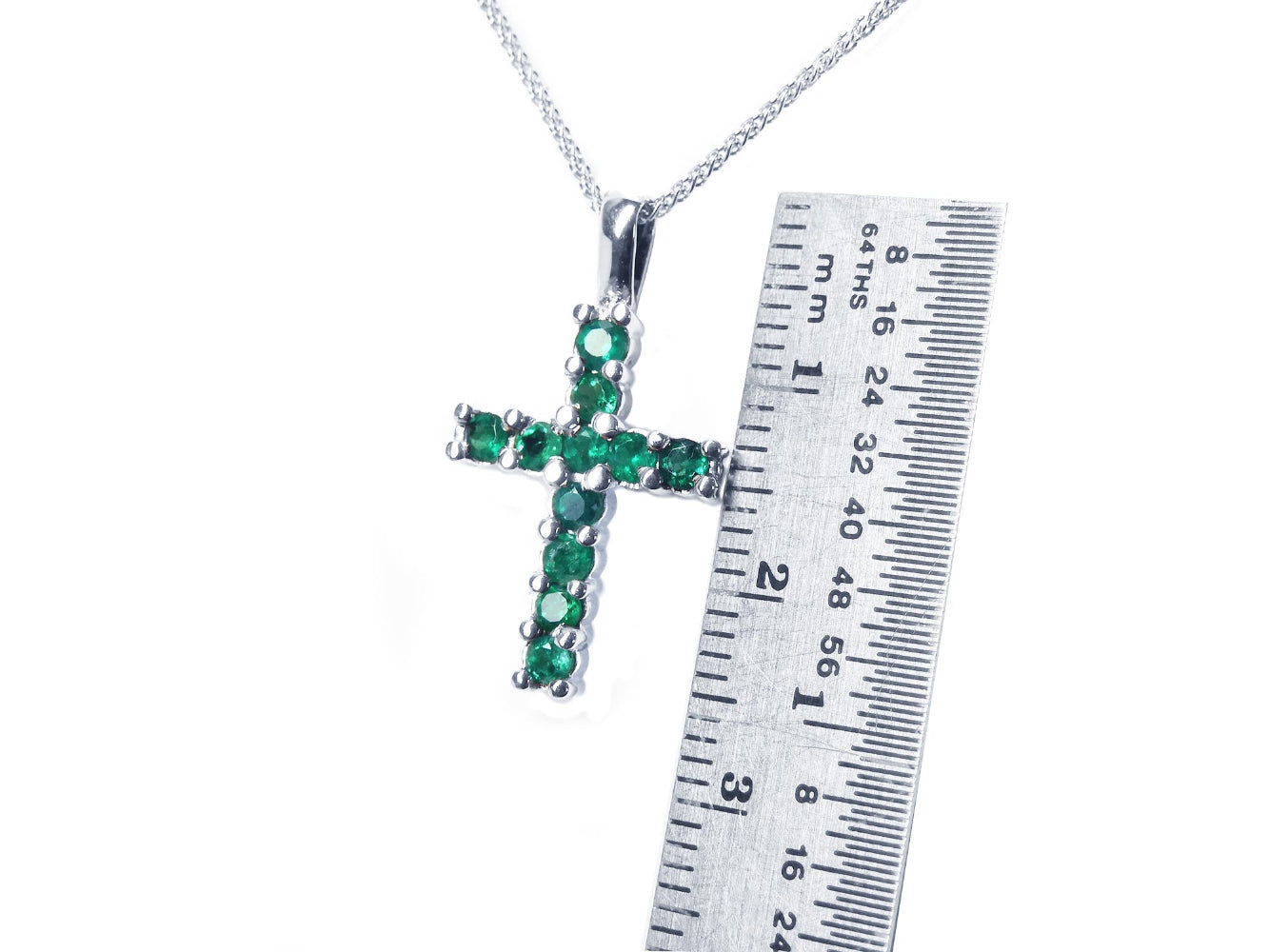 A 18k white gold cross pendant with round cut natural emeralds, next to a ruler for size reference.