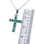 A 18k white gold cross pendant with round cut natural emeralds, next to a ruler for size reference.