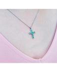 Emerald cross pendant necklace with round-cut emeralds in 18K white gold, worn on a pink top.