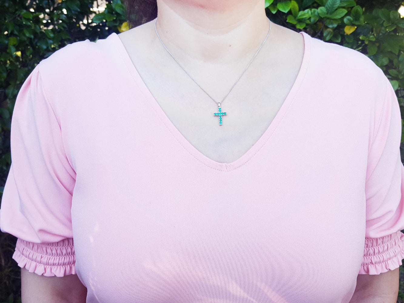 A woman wearing a pink top with a necklace featuring a natural emerald pendant against a blurred green background.