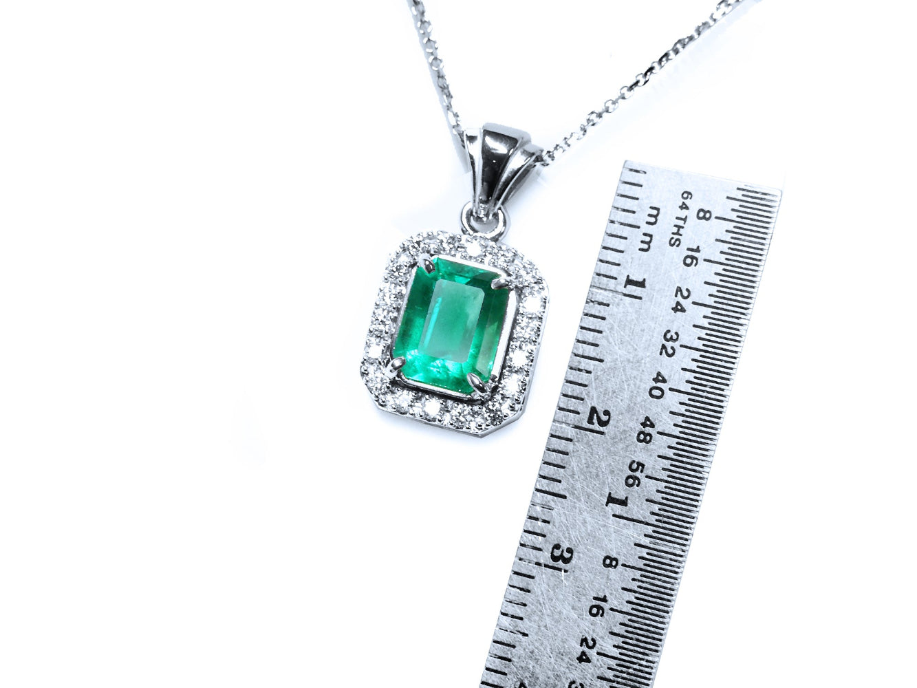Emerald gemstone pendant on a white gold chain with a ruler for scale on a white background.