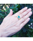 Hand wearing a mens gold ring with a rectangular emerald against a leafy background.
