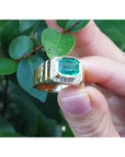 Gold ring with a square natural colombian emerald held by a hand against a leafy background.